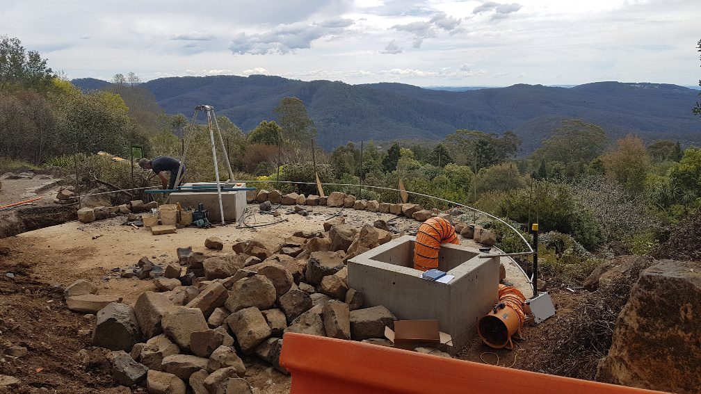 Construction site on a hillside with rocky terrain. A worker is near concrete structures surrounded by rocks. Orange ventilation ducts are placed around. Trees and mountains are in the background under a cloudy sky.