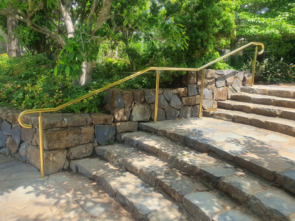 Stone steps with a golden handrail, bordered by a low stone wall and lush green foliage. Sunlight filters through the trees, creating a serene, natural setting.