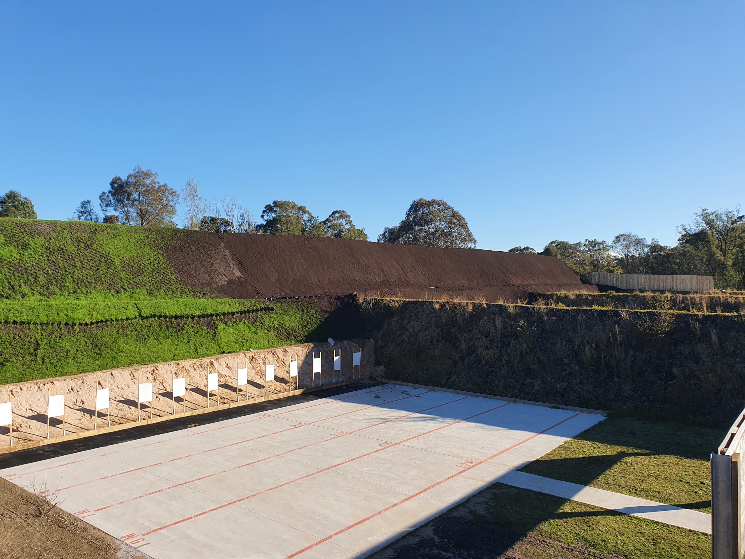 Outdoor shooting range with a concrete firing line and multiple target stands in the distance. The area is surrounded by earthen berms and greenery under a clear blue sky.