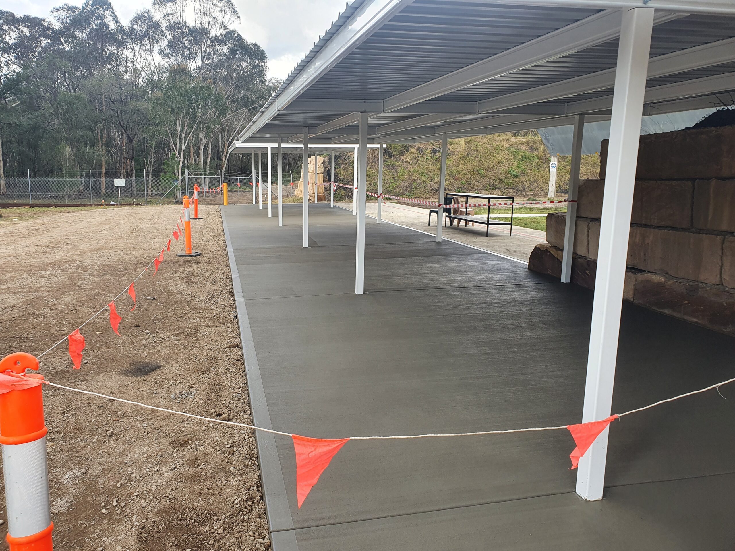 A newly constructed covered walkway with a smooth concrete surface. Red flags on a rope and orange safety cones line one side. Trees and a dirt area are visible in the background.