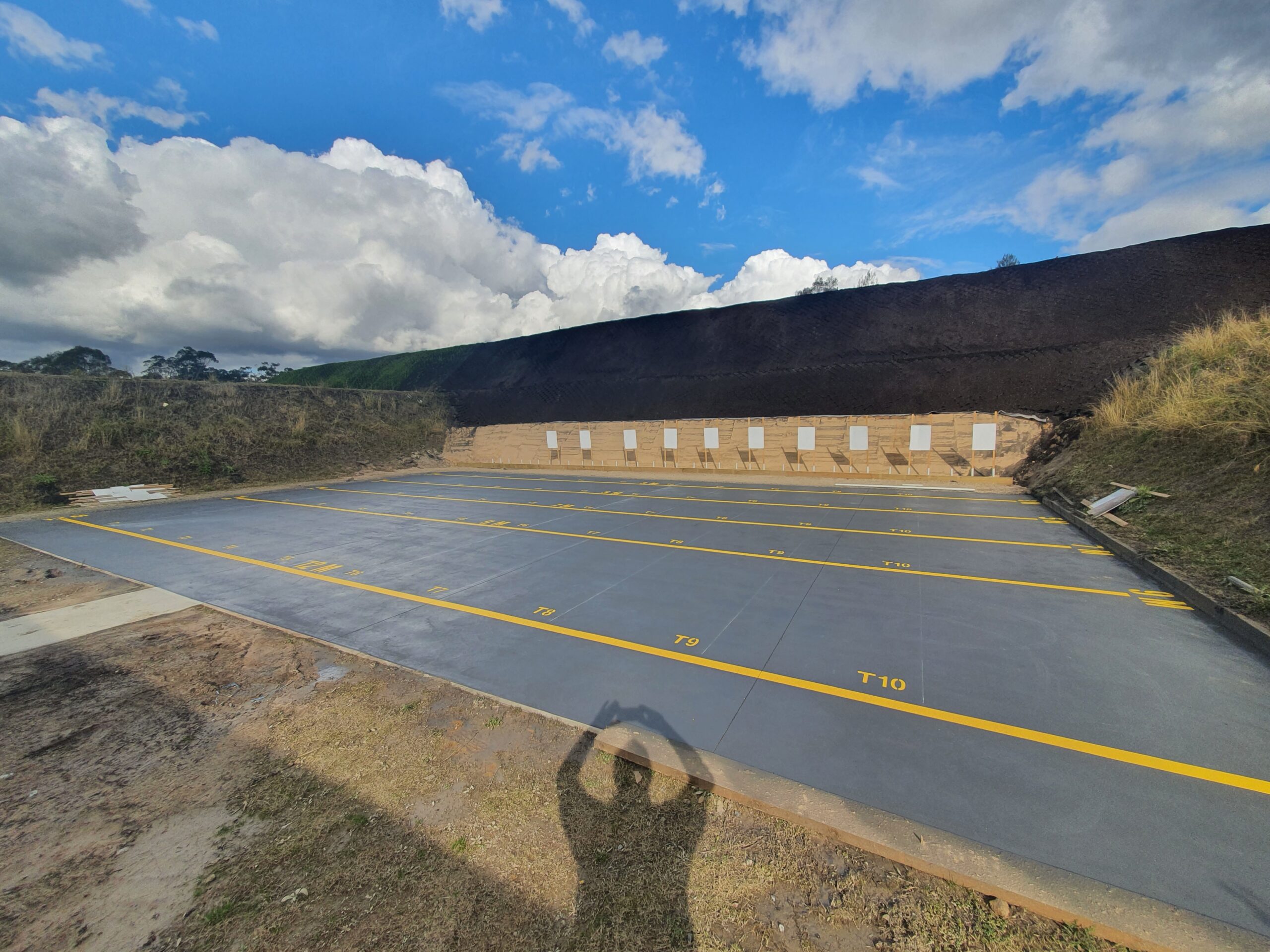 A large, empty outdoor shooting range with numbered lanes painted in yellow on a gray concrete surface. It's surrounded by earth embankments, with a person casting a shadow in the foreground, holding up a camera. The sky is partly cloudy.