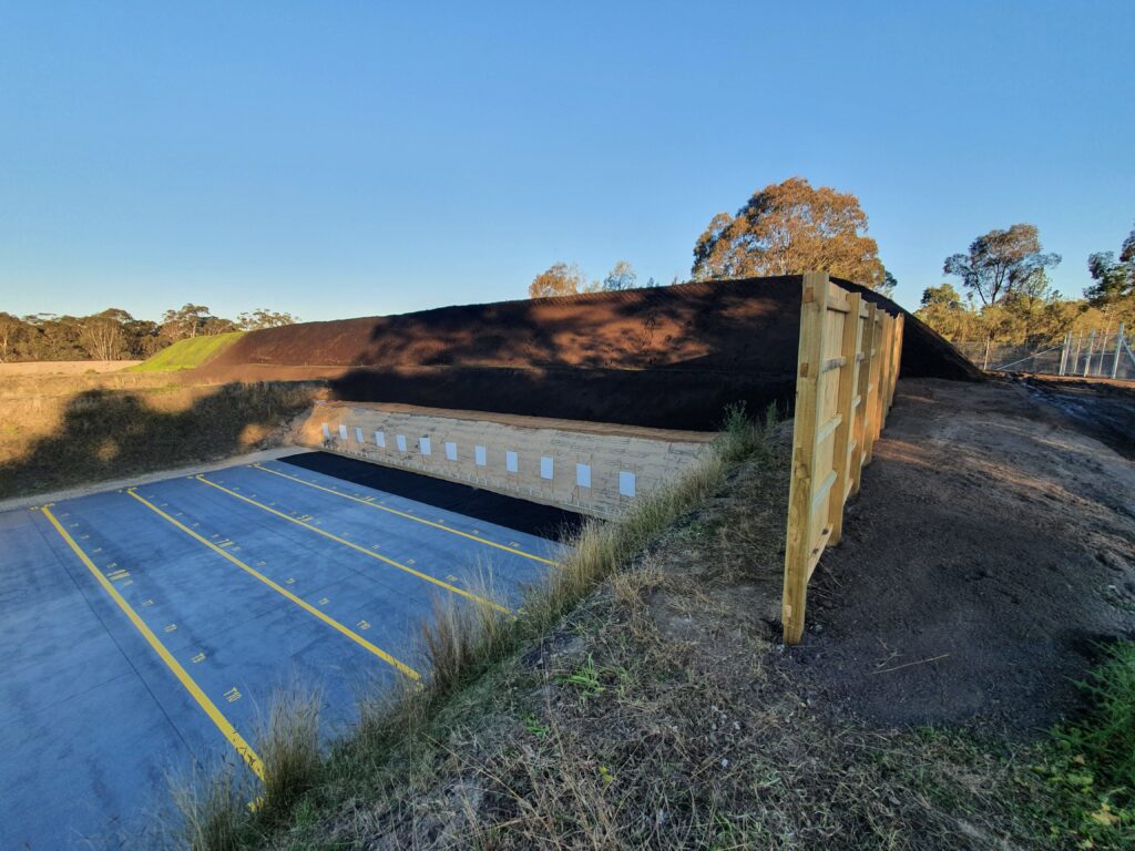 A wooden barrier and mound of soil border a parking lot. The lot features numbered spaces and yellow outlines, adjacent to a tree line under a clear blue sky.