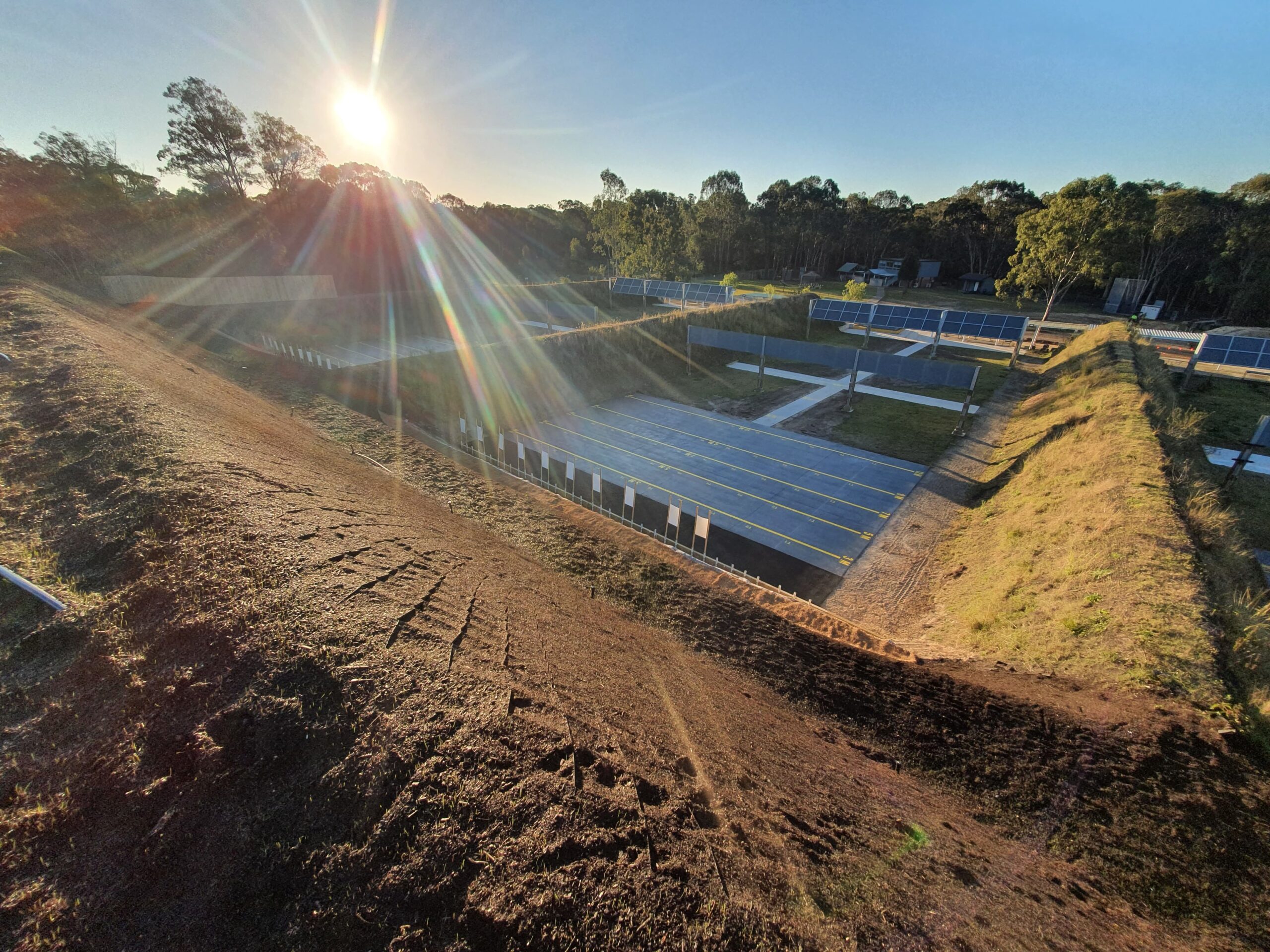 Sunlight beams over a large, fenced solar farm set within grassy, earth-bordered terraces. Rows of solar panels are aligned systematically, surrounded by trees under a clear blue sky.