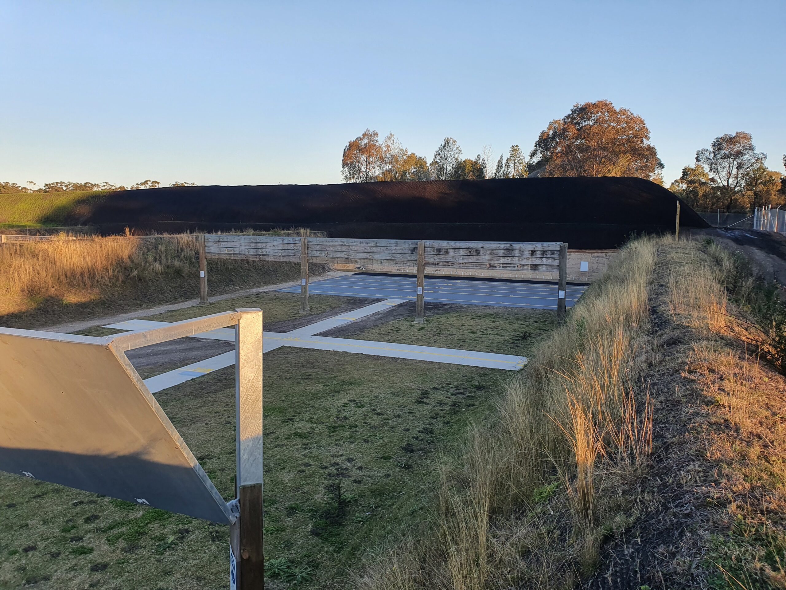 A shooting range featuring several concrete shooting lanes with wooden barriers. The area is surrounded by grassy fields and bushes, and a dark backstop is visible in the distance. Trees and a clear sky are in the background.