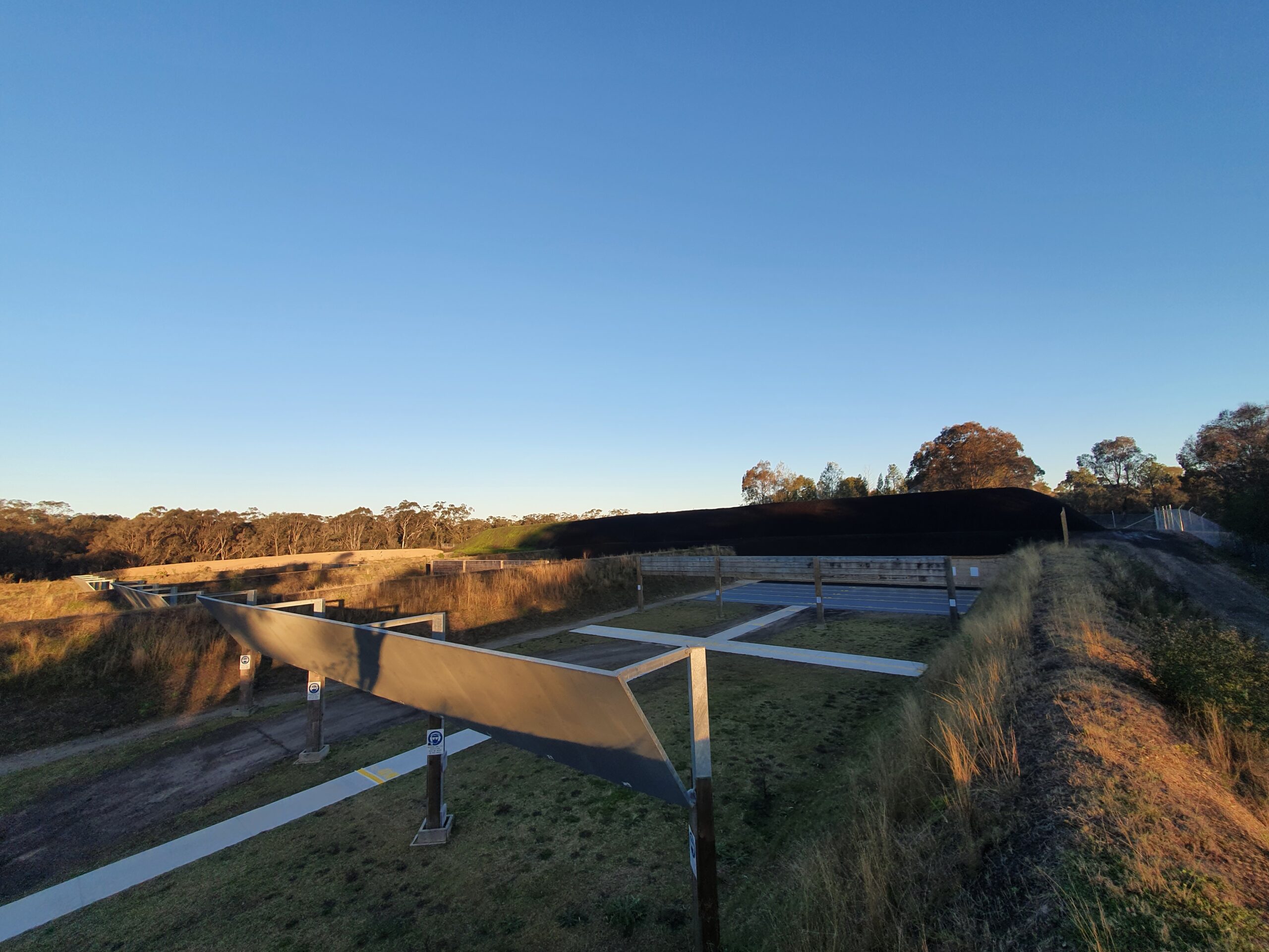 Outdoor firing range with several shooting lanes under clear blue sky. The lanes are lined with concrete paths and have angled metal shelters. Surrounding areas are grassy with trees in the background, under late afternoon sunlight.