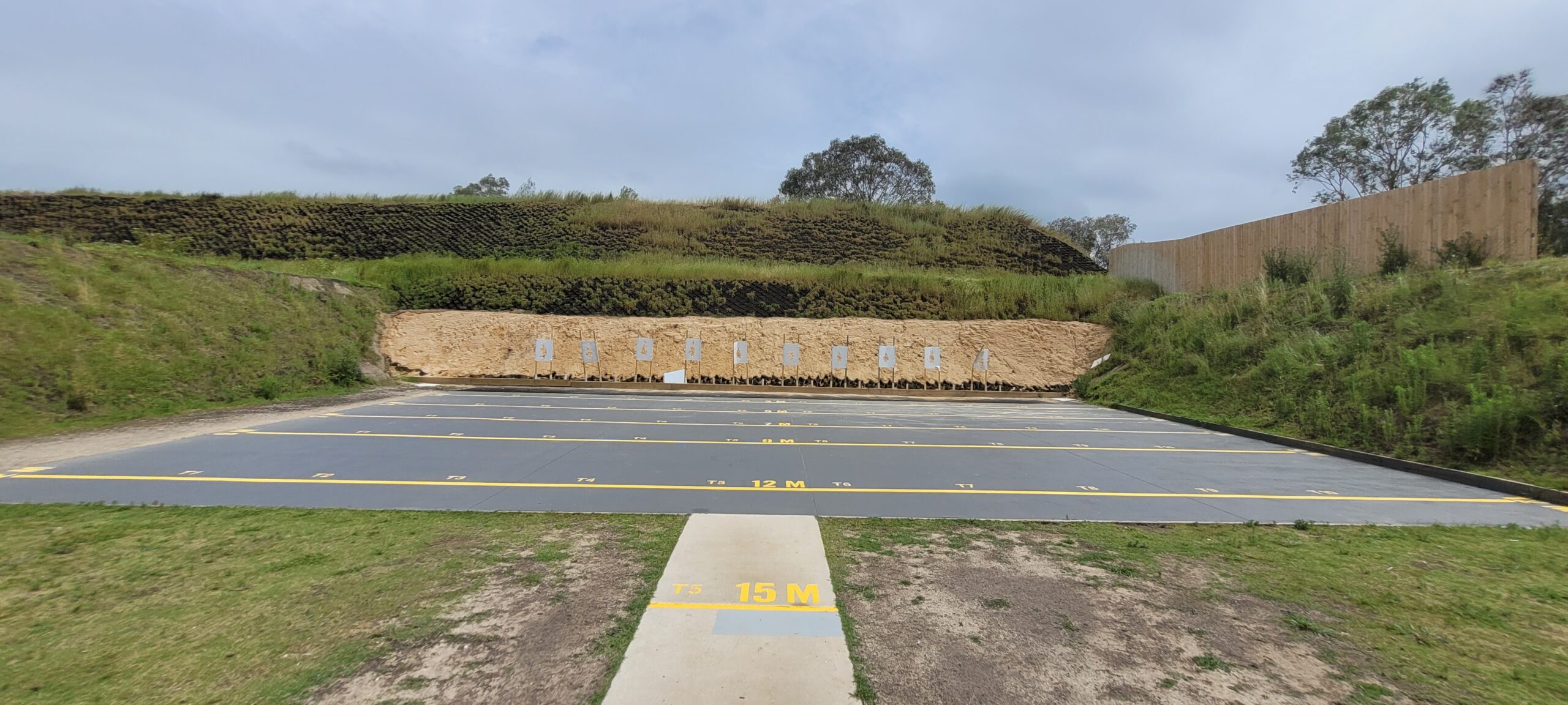 A shooting range with multiple target sheets in the background. Marked distances of 12 and 15 meters are visible on the ground. The range is surrounded by grassy hills and a cloudy sky overhead.