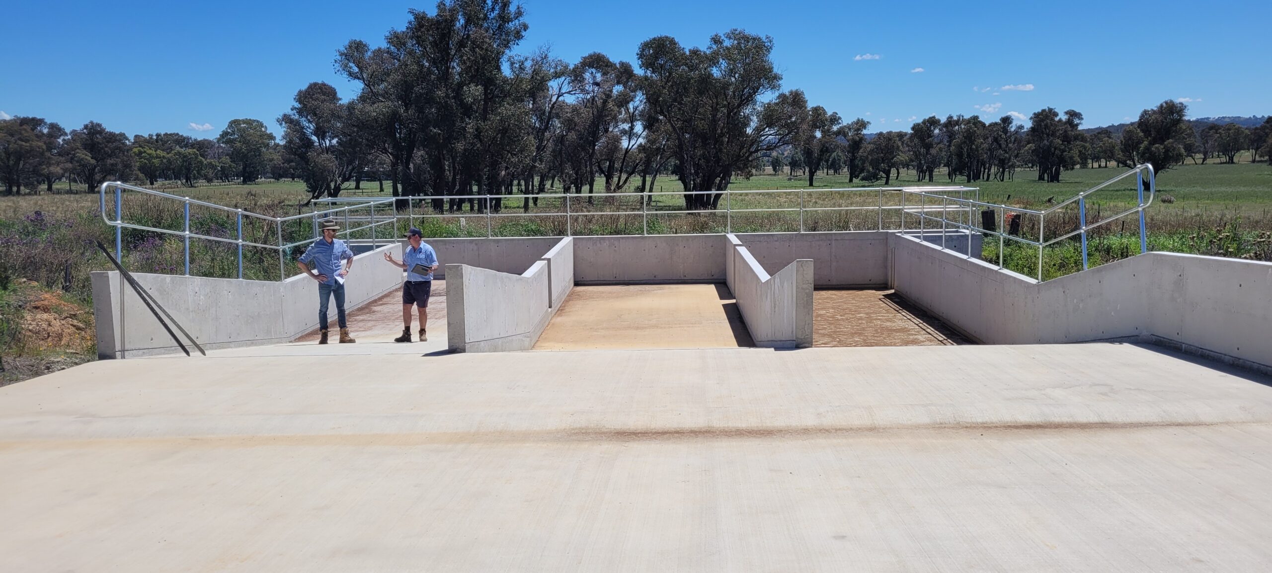 Two people stand on a large concrete structure with railings, possibly a water management or flood control installation. Trees and grassy fields are in the background under a clear blue sky.