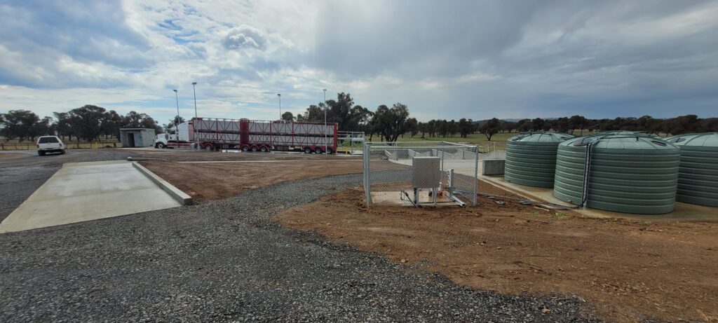 A rural site with large green water tanks, a fenced utility area, and a parked truck with red trailers. The landscape includes gravel and dirt areas, with scattered trees in the background under a cloudy sky.