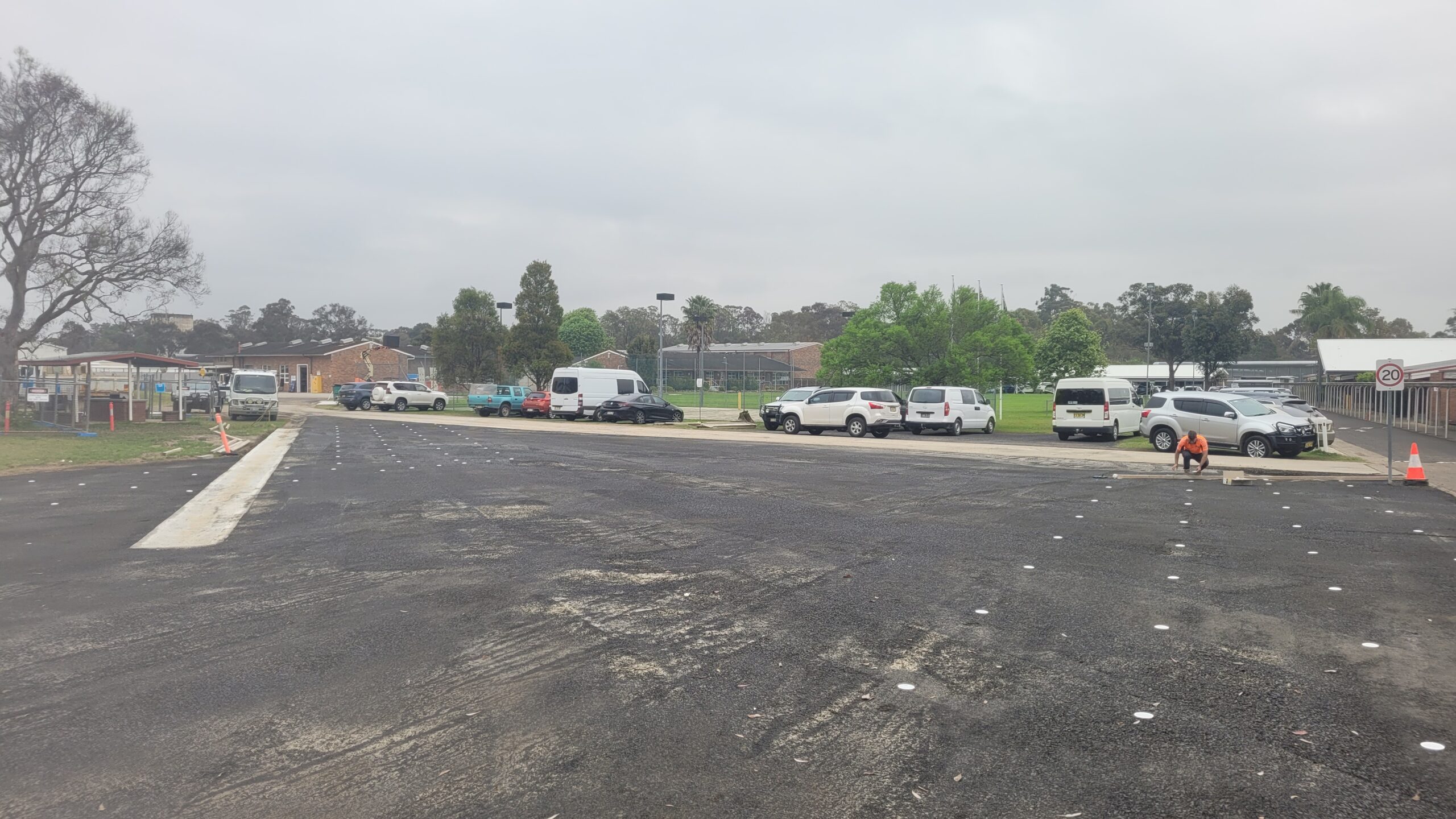 A parking lot with several vehicles parked, including white vans and cars. The ground is paved with dark asphalt, and the area is bordered by trees and buildings in the background under a cloudy sky. A person walks a dog on the right.