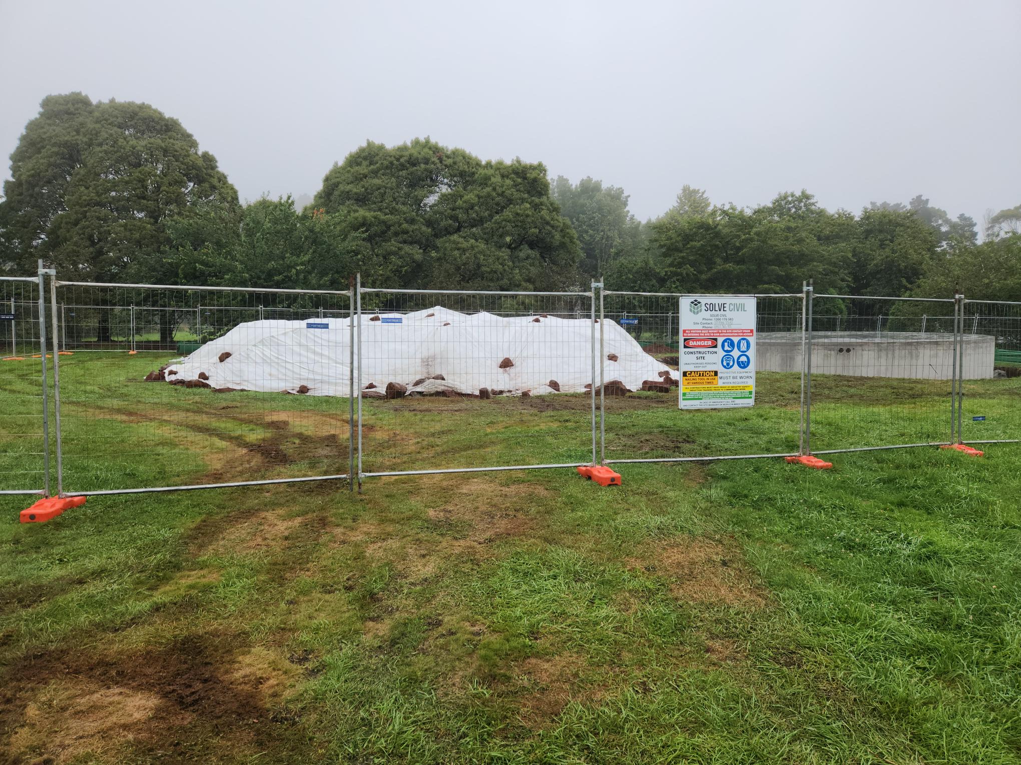 A fenced-off area in a grassy field contains a covered pile of white material. Surrounding it is a metal fence with a sign featuring safety and site information. Trees are visible in the misty background.