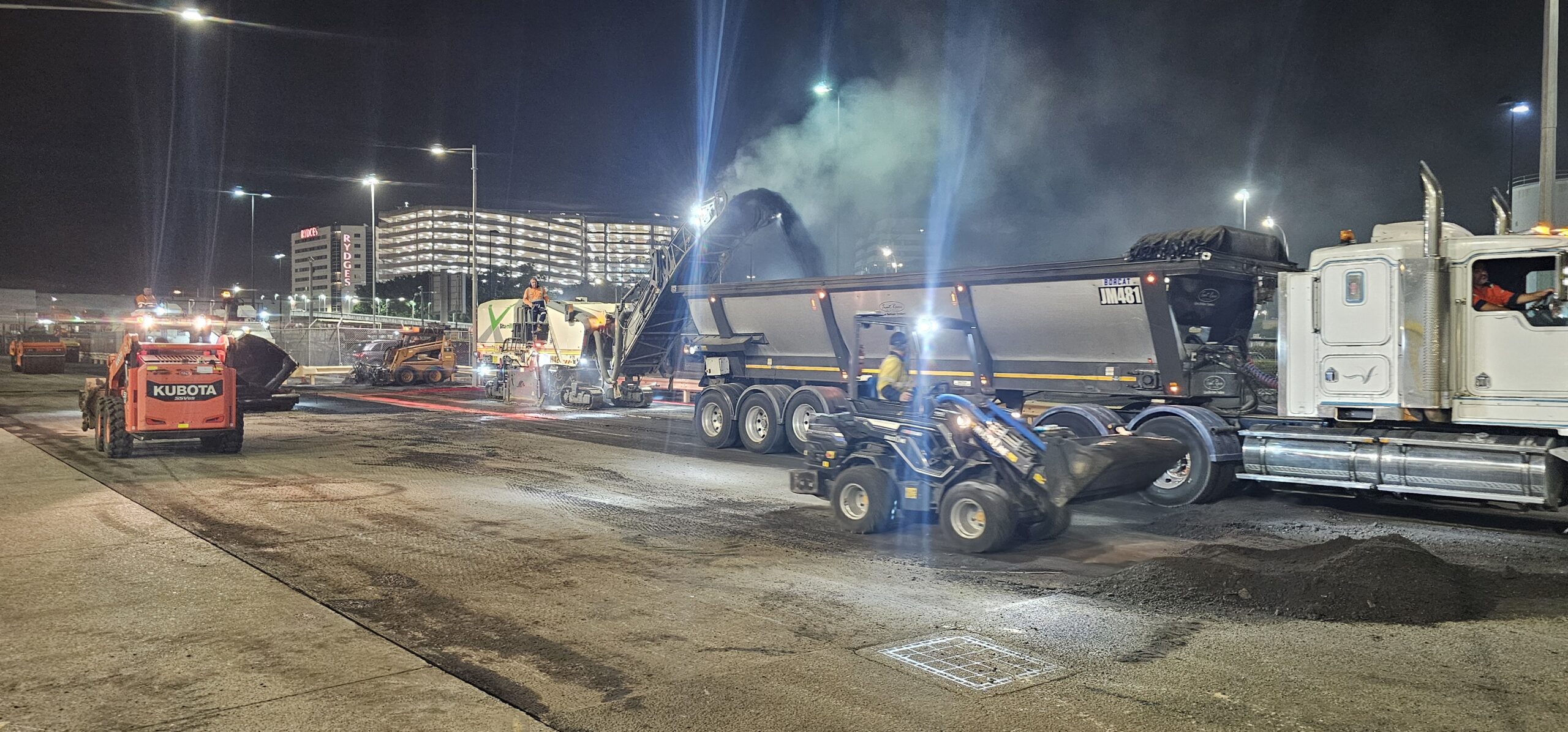 Construction workers repaving a road at night under bright lights. Heavy machinery and trucks are visible, with steam rising from the freshly laid asphalt. A modern building in the background is illuminated.