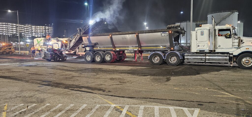 Nighttime construction scene with a large truck alongside a machine offloading materials into it. Workers in high-visibility clothing are present, and a building with bright lights is visible in the background.