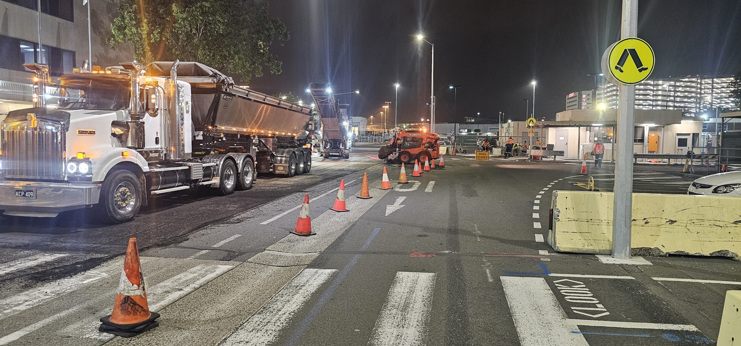 Nighttime construction scene with trucks and equipment on a road. Orange cones line the street, directing traffic. A construction vehicle operates nearby under streetlights. Buildings and a pedestrian crossing sign are visible in the background.