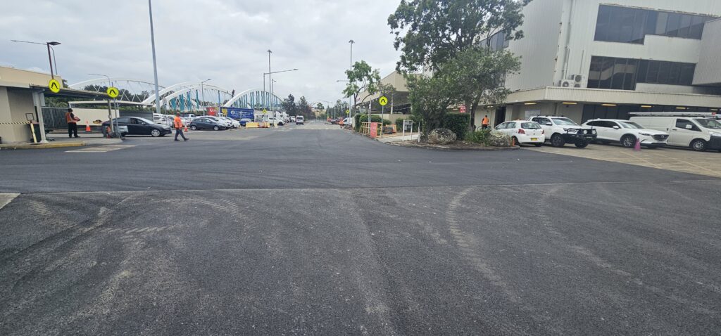 Wide empty street view, bordered by office buildings and parked cars. Overcast skies and construction materials visible on the left. Pedestrians are walking along the sidewalk. Arched bridge is in the distance under cloudy skies.