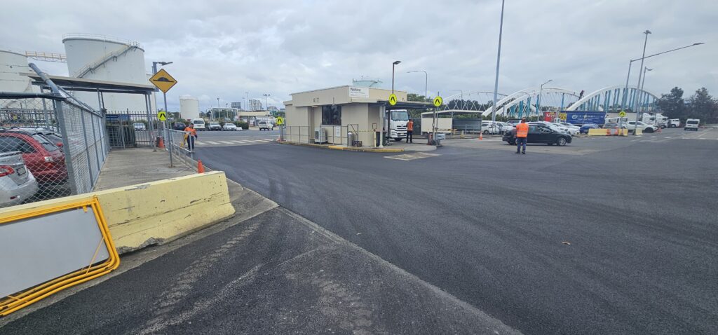 Industrial site with an asphalt road and a small building in the center. A truck is parked at the building. Several people in orange vests stand nearby. Cars are parked on the left, and a large bridge is visible in the background under a cloudy sky.