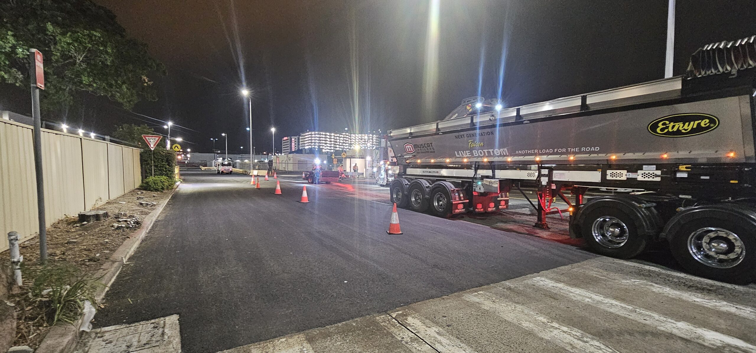 Night scene depicting a street with traffic cones and a large truck parked on the right. Streetlights illuminate the area, with a building visible in the background. A yield sign is seen to the left, and the road is partially closed off.