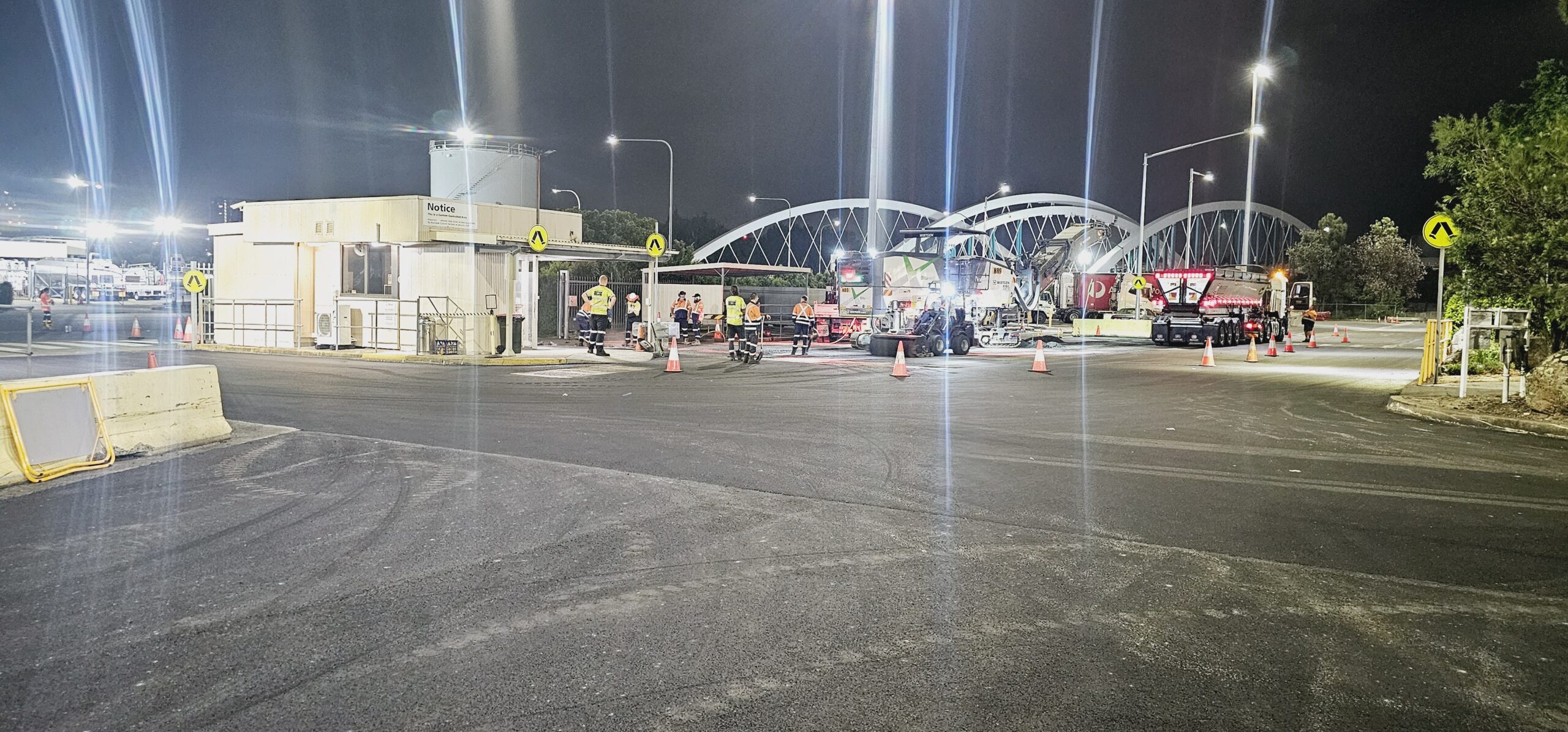 Nighttime road construction scene with workers in high-visibility clothing operating machinery. Traffic cones line the area, and bright lights illuminate the site. A distinctive arched bridge is visible in the background.