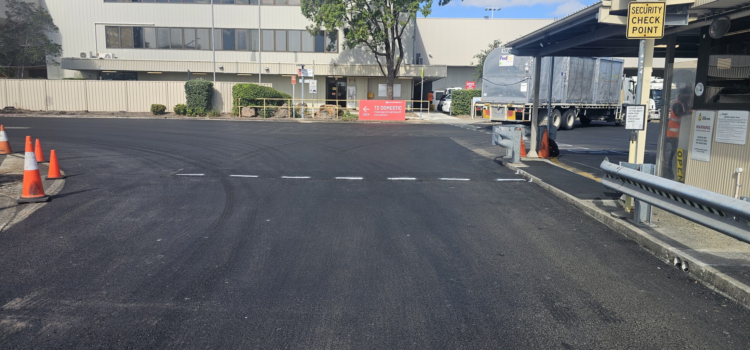 Paved roadway at a security checkpoint with traffic cones on the left side. A building and parked truck are visible in the background. A red sign is placed near the building. The area appears to be an industrial or commercial zone.