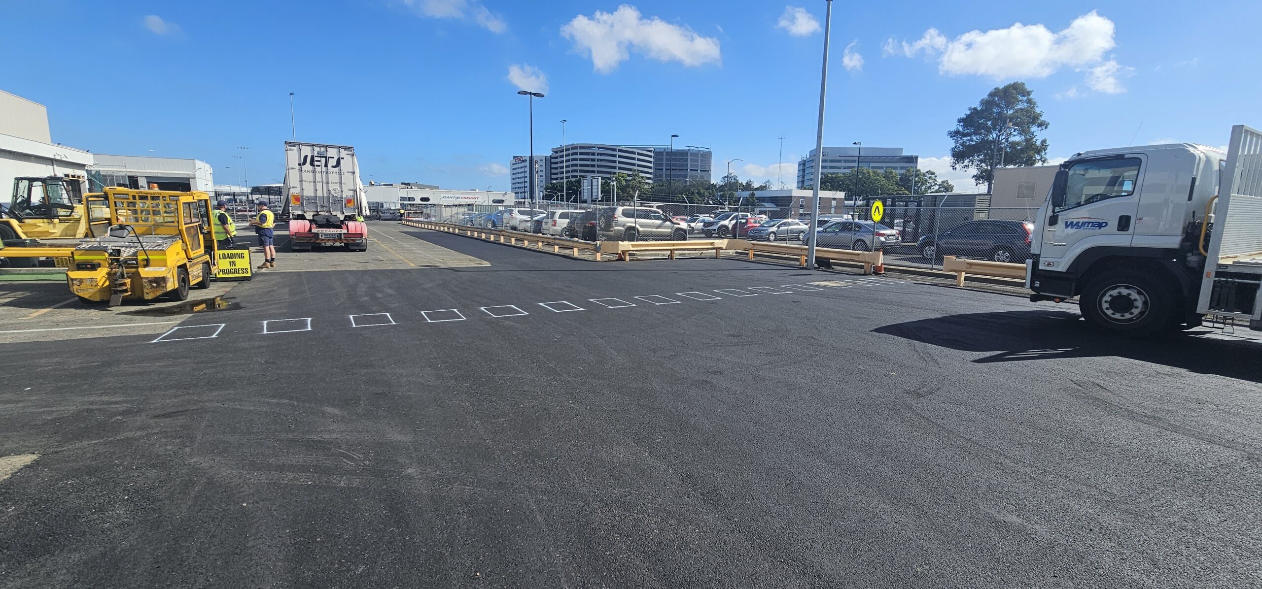 Parking lot with freshly paved surface under a clear blue sky. Yellow and white industrial vehicles are parked to the left. Cars are parked in the background, with tall buildings visible. Two large white trucks are on the right.