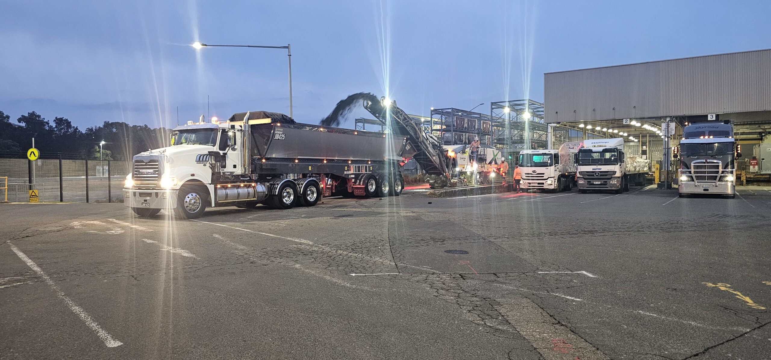 A line of trucks is parked at an industrial site in the evening. Bright lights illuminate the area, with trucks staged under a large roofed structure. One truck is actively being loaded with materials. The sky is dim, suggesting dusk or dawn.