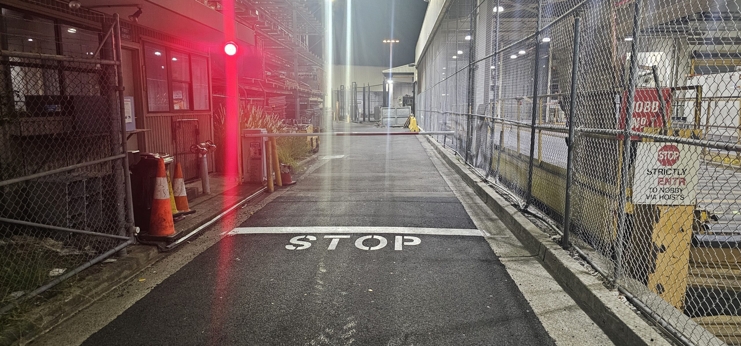 A dimly lit industrial entrance with a red traffic light glowing. A barrier blocks the narrow road, and 