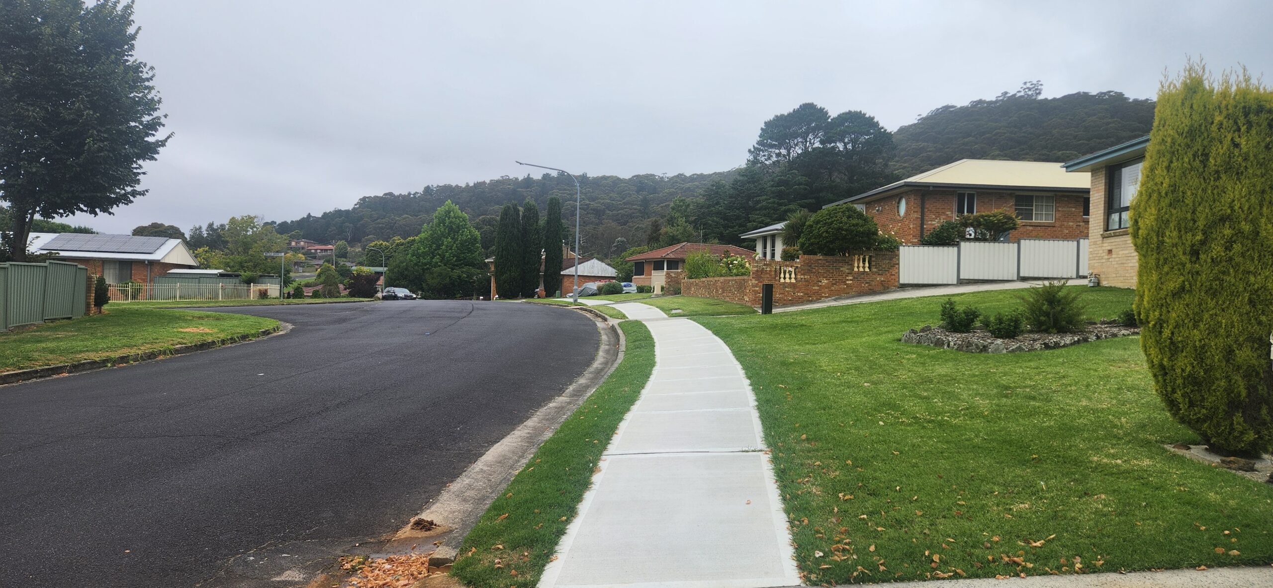 A suburban street curves gently to the left, lined with houses and well-maintained lawns. A concrete sidewalk parallels the road. Trees and shrubs border the area, and wooded hills are visible in the background under a cloudy sky.