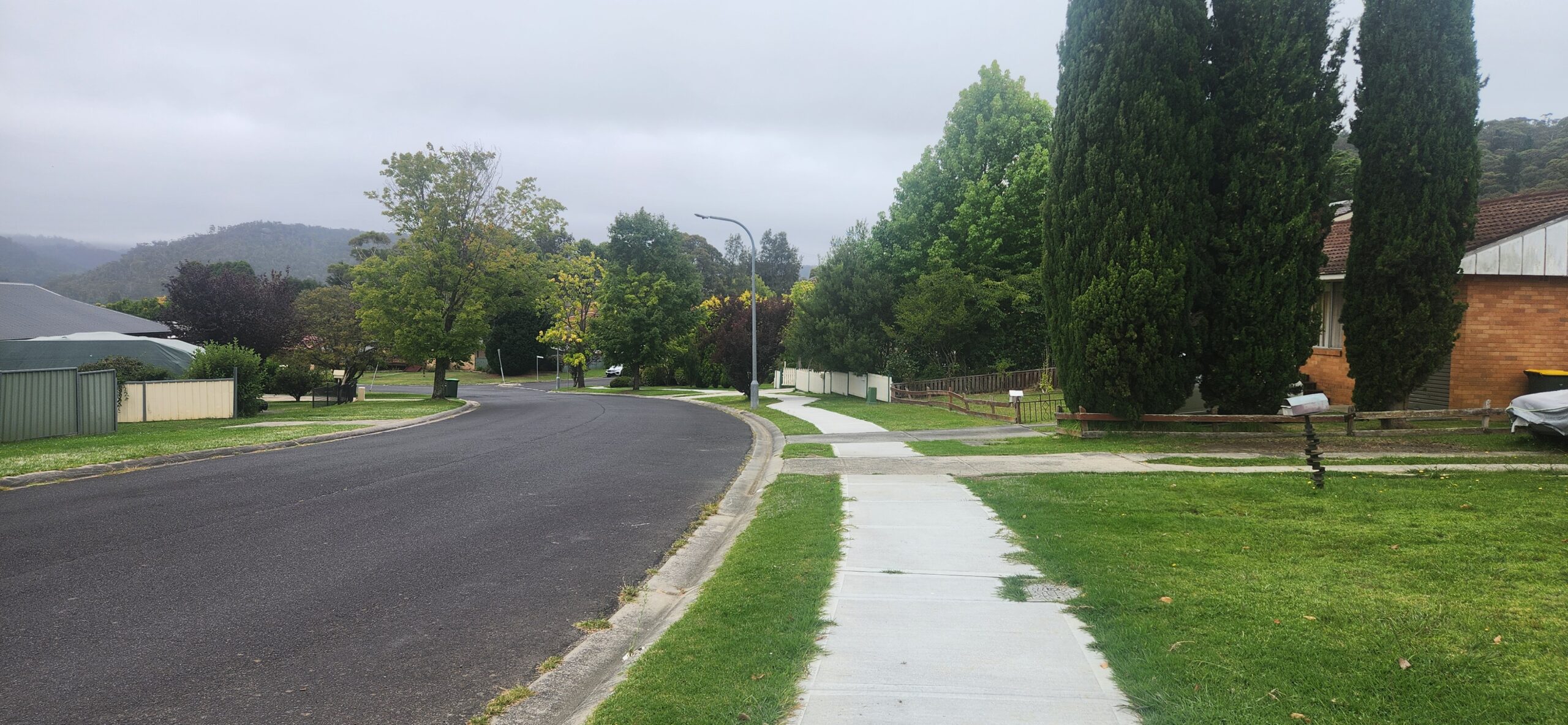 A quiet suburban street with a curved road, lined by grass and sidewalks. Tall green trees are on the right, with houses partially visible. Overcast sky with distant hills in the background.