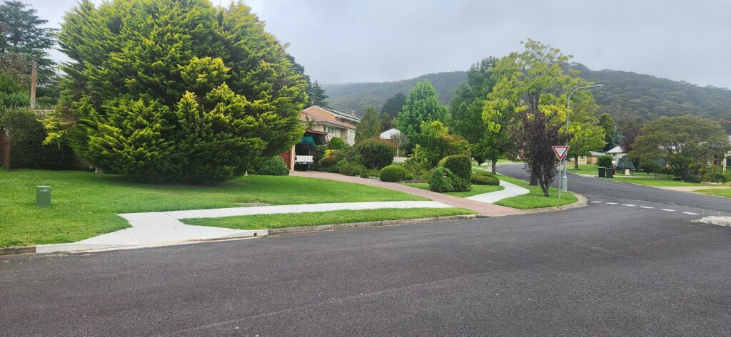 A suburban street with a crossroads, surrounded by lush green trees and houses. The road is empty, and the sky is overcast. Sidewalks intersect at the corner, and the area is well-maintained and verdant. Hills are visible in the background.