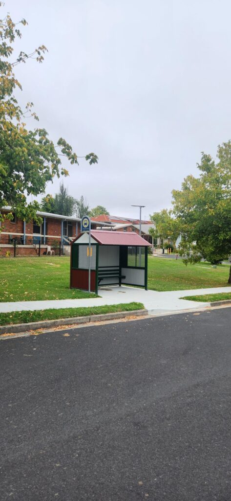 A small, covered bus stop with benches sits beside a quiet street. Surrounding it are well-maintained green lawns and trees. In the background, there are residential houses with brick exteriors. The sky is overcast.