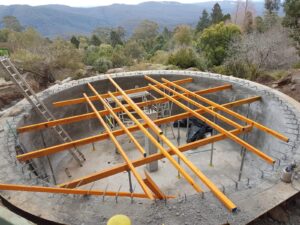 Circular concrete structure under construction, with wooden beams placed across the diameter. Surrounding scenery includes trees and a mountainous background. Ladders are visible, and construction materials are scattered inside the structure.