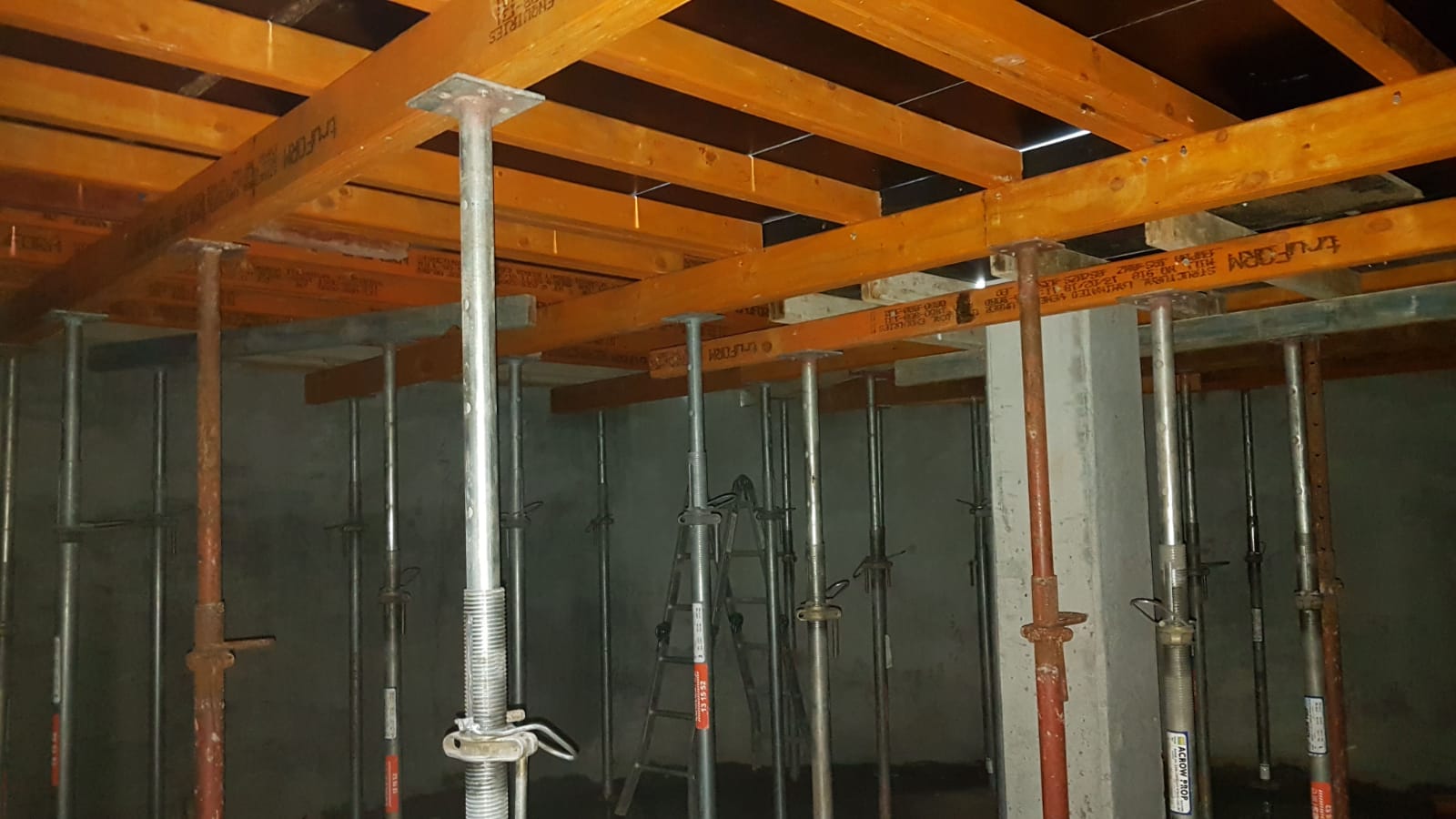 Construction site interior with metal supports and wooden beams. Several adjustable steel props are arranged vertically to hold the overhead framework. The setting appears dimly lit, showing structural elements in a partially built area.