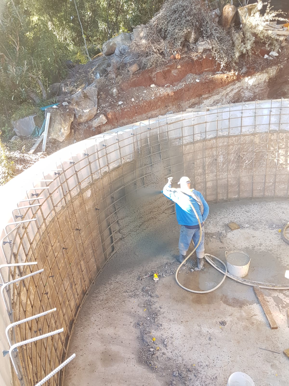 A worker in a blue outfit sprays concrete on the interior of a partially constructed pool, surrounded by rebar. Construction materials are scattered around the site, with a dirt hill and trees in the background.