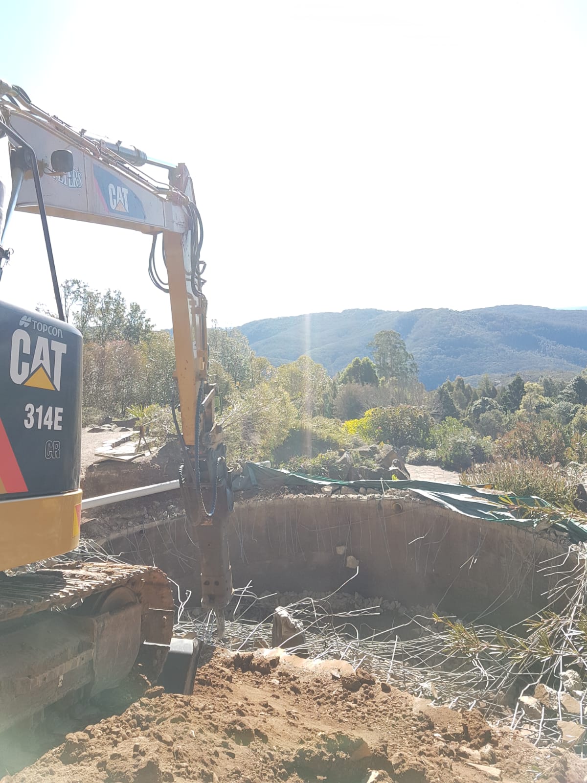 An excavator works on a construction site with rubble and debris in the foreground. Trees and hills are visible in the background under a clear sky.