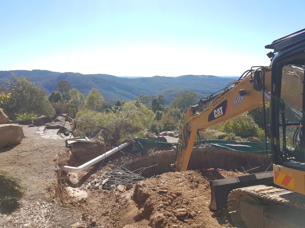 An excavator is digging earth on a hillside with a view of green mountains and blue sky in the background. A large pipe is partially buried in the ground, surrounded by dirt and rocks. Vegetation lines the pathway nearby.