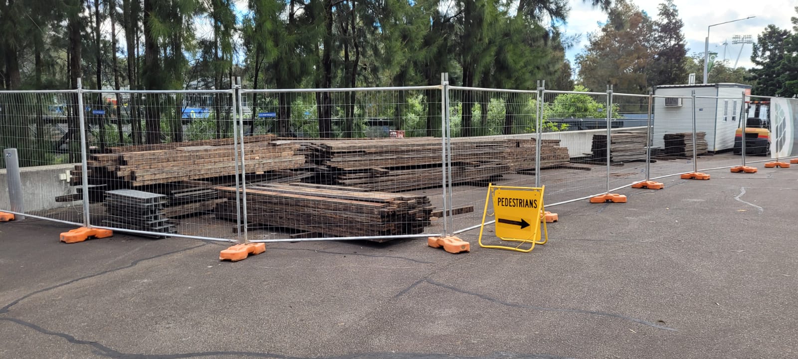 A construction site with stacks of wooden planks behind a metal fence. An orange and yellow pedestrian sign with an arrow points right. Trees and a small building are in the background.