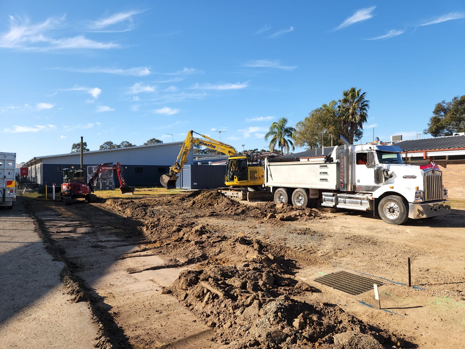 Construction site with a white dump truck, a yellow excavator, and a red earthmover. The ground is dug up, and there are buildings and palm trees in the background under a clear blue sky.