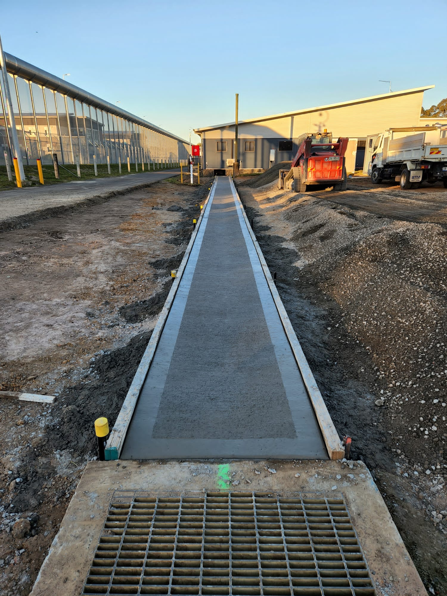 A freshly paved concrete sidewalk stretches between piles of dirt. Construction vehicles and equipment are visible in the background near a building. The area is under clear blue skies.