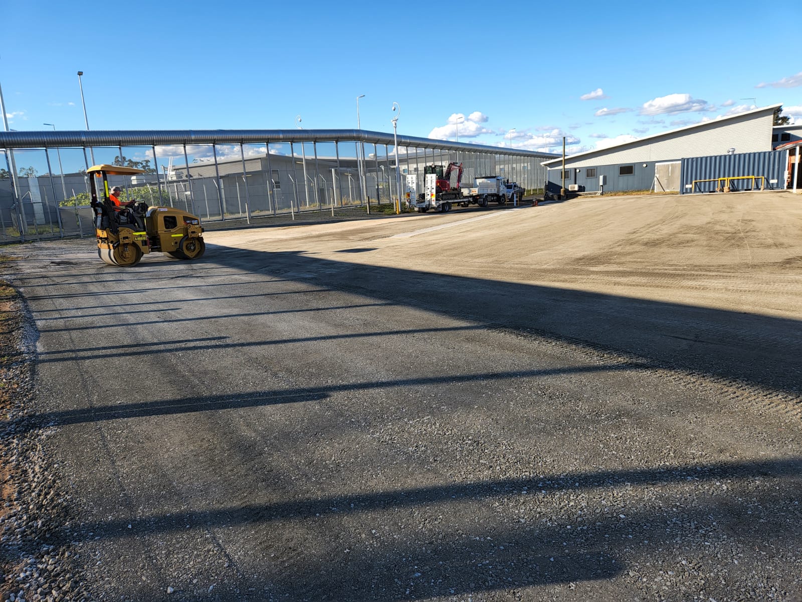 A construction site with a compacted dirt area and several buildings in the background. A road roller is working on the surface, and another vehicle is parked nearby. The sky is clear with a few clouds.