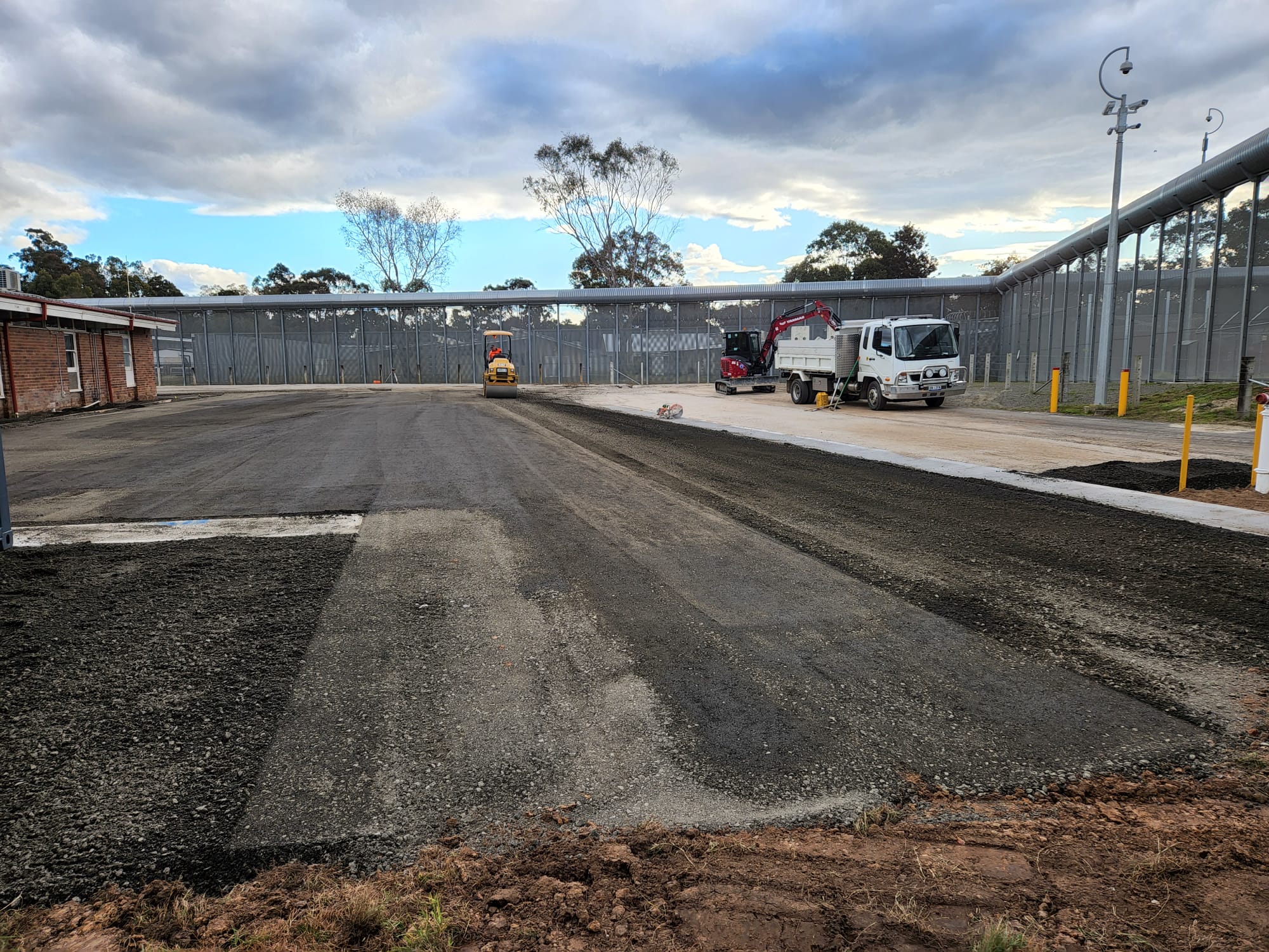 A construction site with fresh asphalt paving and machinery. A truck and a steamroller are parked on the right side, under a partly cloudy sky. A metallic fence and some trees are visible in the background.