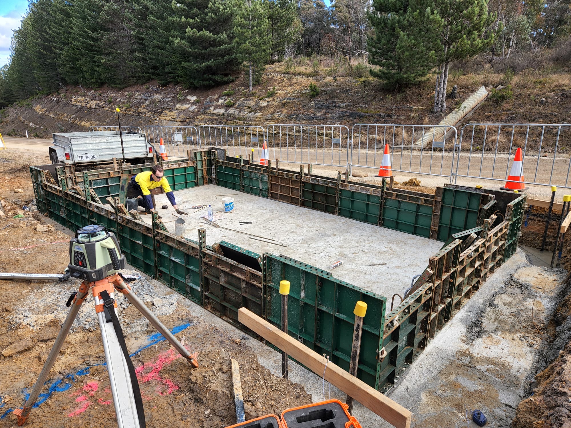 Construction worker inside a rectangular pit surrounded by green formwork panels. The area is bordered by orange cones and a metal fence. A truck is parked nearby, and there's surveying equipment in the foreground. Trees line the roadside.