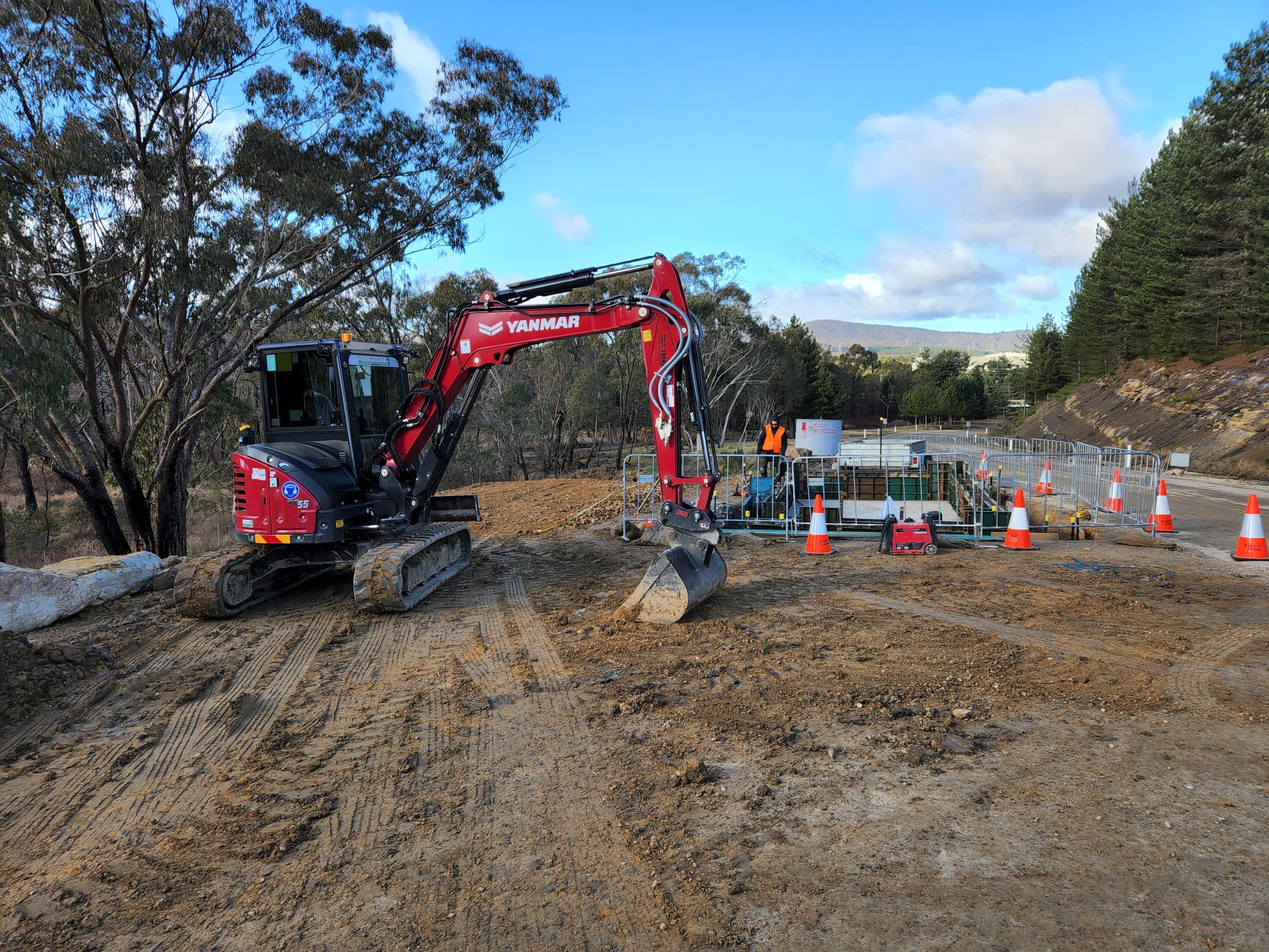 A red excavator is parked on a dirt construction site surrounded by orange traffic cones. Workers in safety gear are visible in the background near metal fencing. Trees and a blue sky are in the distance.