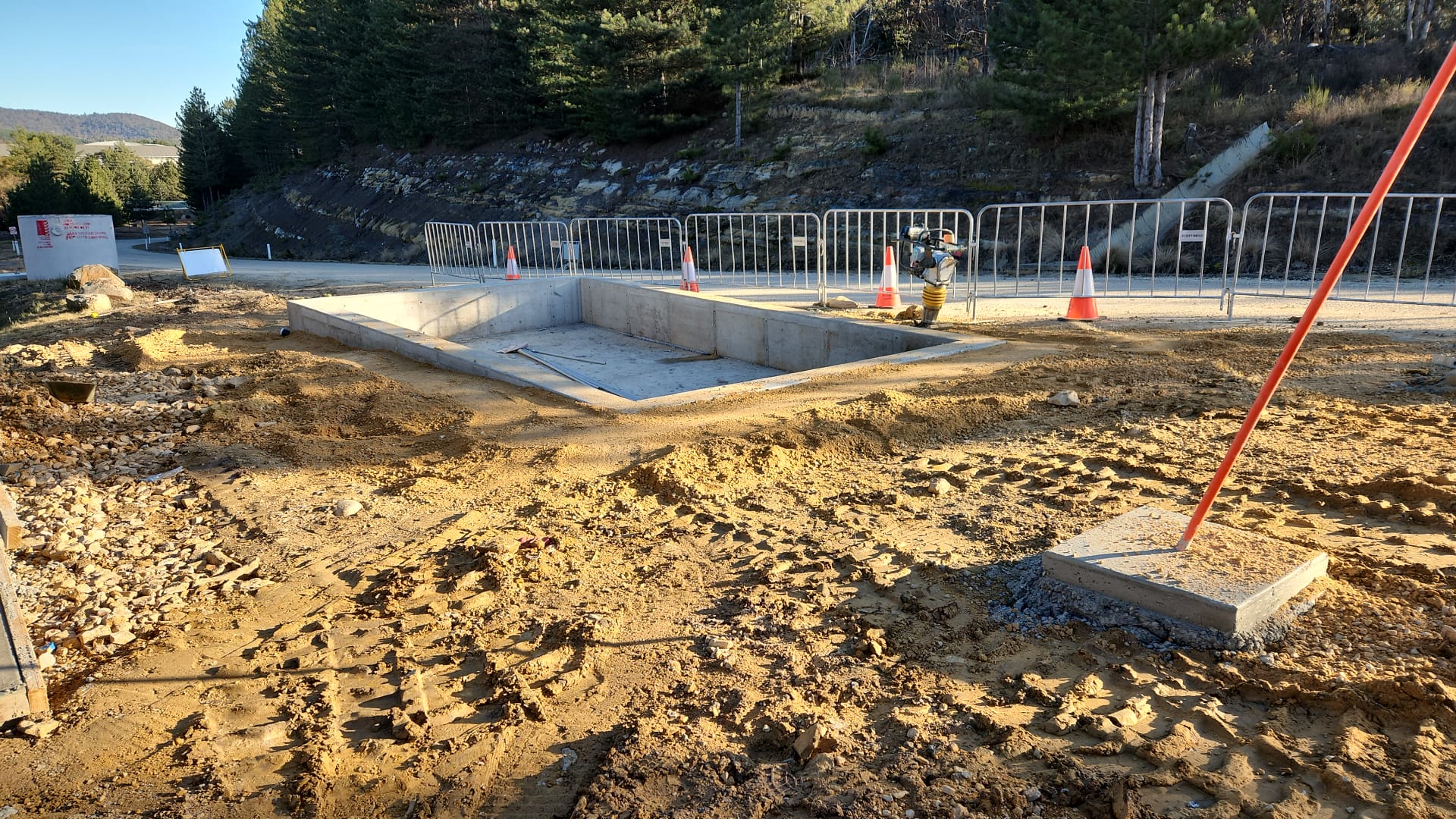 A construction site with a rectangular concrete foundation in progress, surrounded by dirt and tire tracks. Orange safety cones and a metal fence encircle the area. Trees and hills are visible in the background under a clear sky.