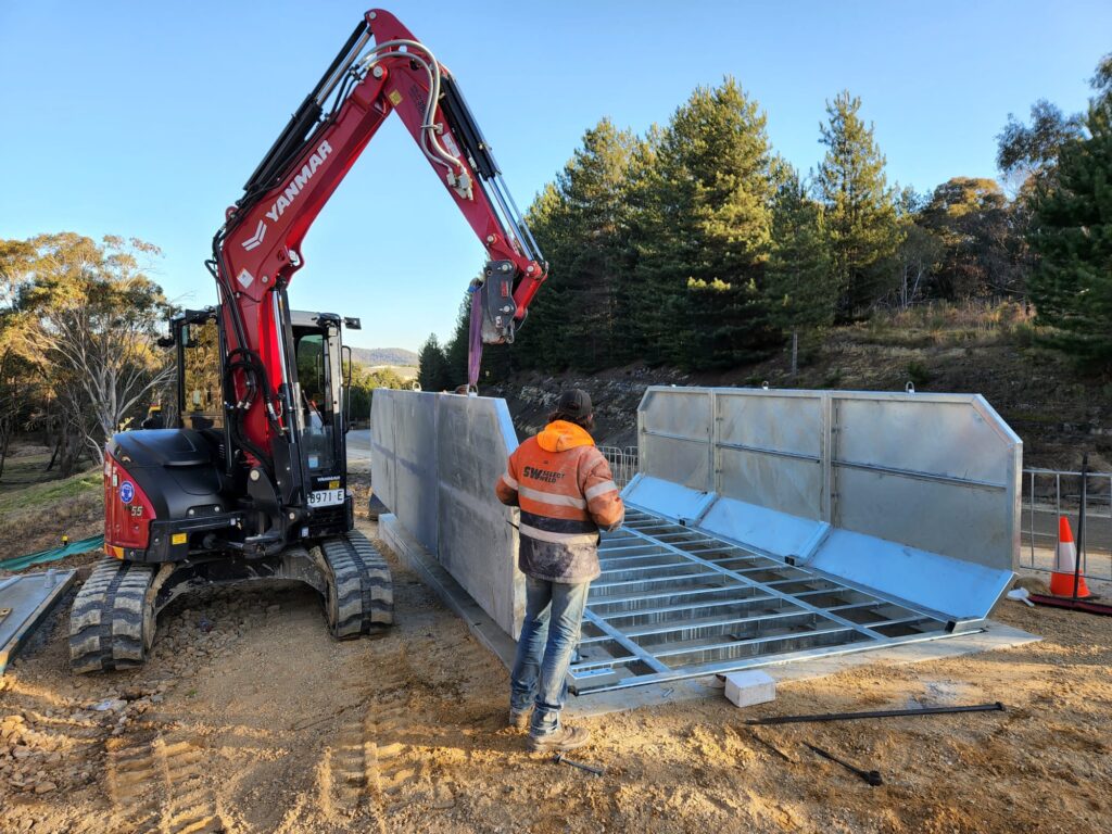 A construction worker in an orange and black jacket operates a red excavator, maneuvering a large metal structure. The scene is outdoors, surrounded by trees and dirt, with traffic cones visible in the background.