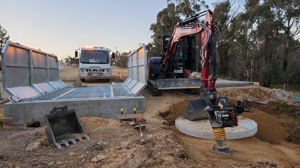 Construction site with a small red excavator and a white truck on a dirt road. Metal structures, possibly trench boxes, are being installed. Surrounded by trees, the site has various construction equipment and materials scattered around.