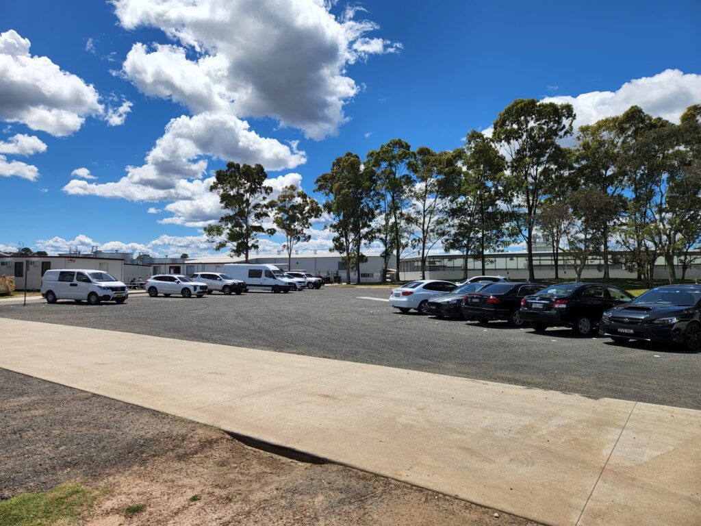 A parking lot with several cars and a white van under a bright blue sky with scattered clouds. Tall trees line the background, and a pathway runs along the foreground.