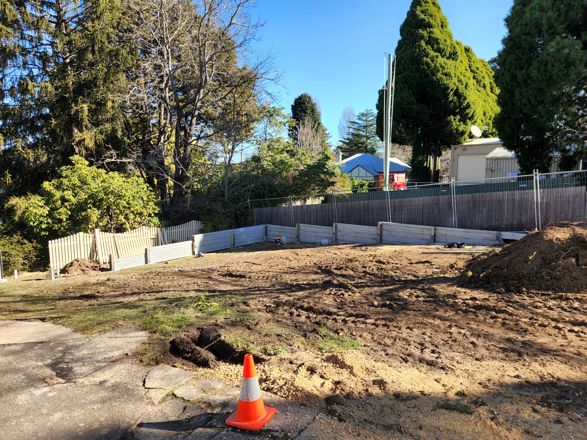 A dirt construction site with a fenced perimeter and scattered wooden planks. A single orange traffic cone sits at the edge. Trees and a house are visible in the background under a clear blue sky.