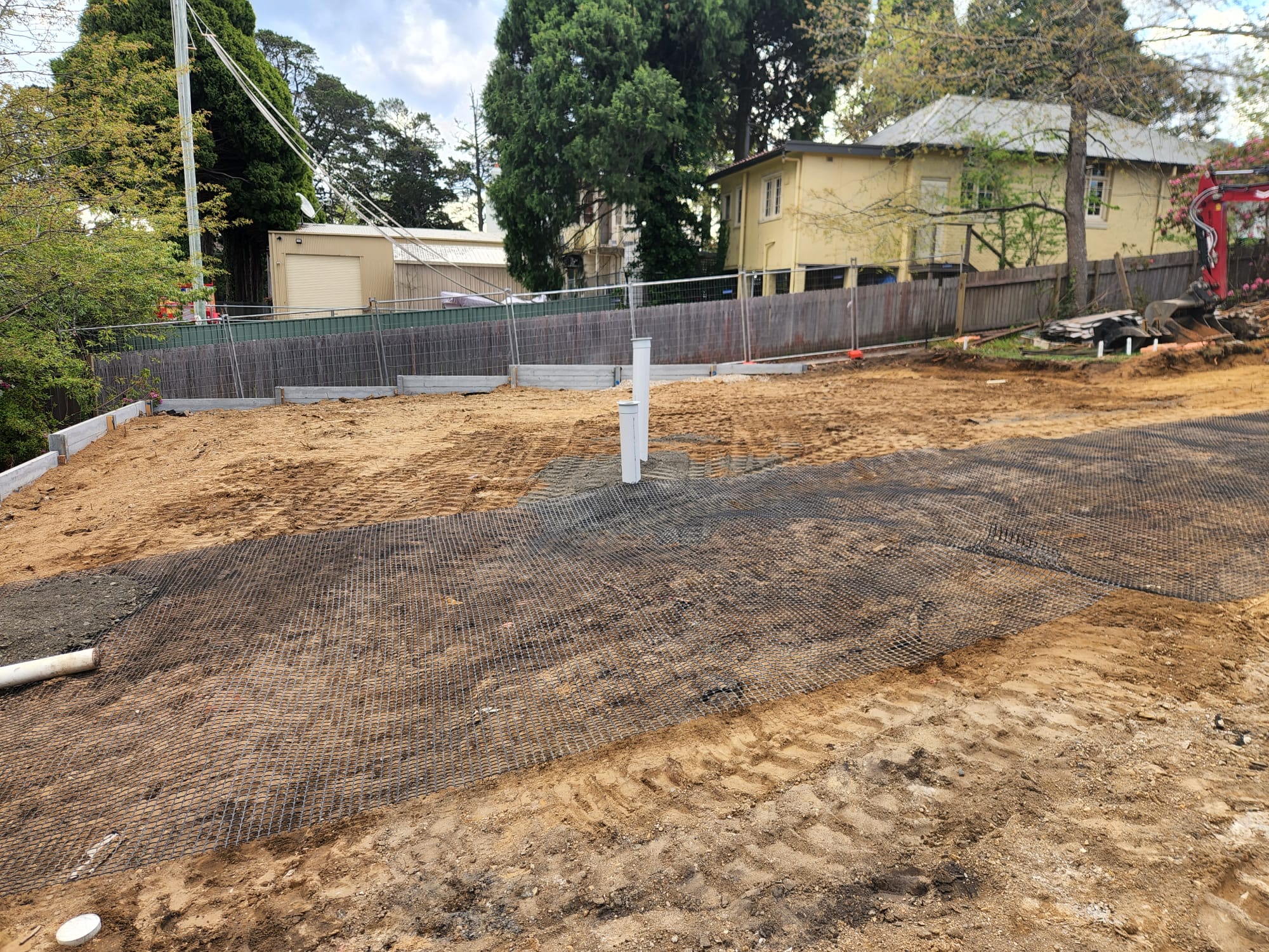 Construction site with a cleared, leveled dirt area and protective netting on the ground. There's a white pipe standing vertically in the center. Trees, a metal fence, and a yellow building are visible in the background.