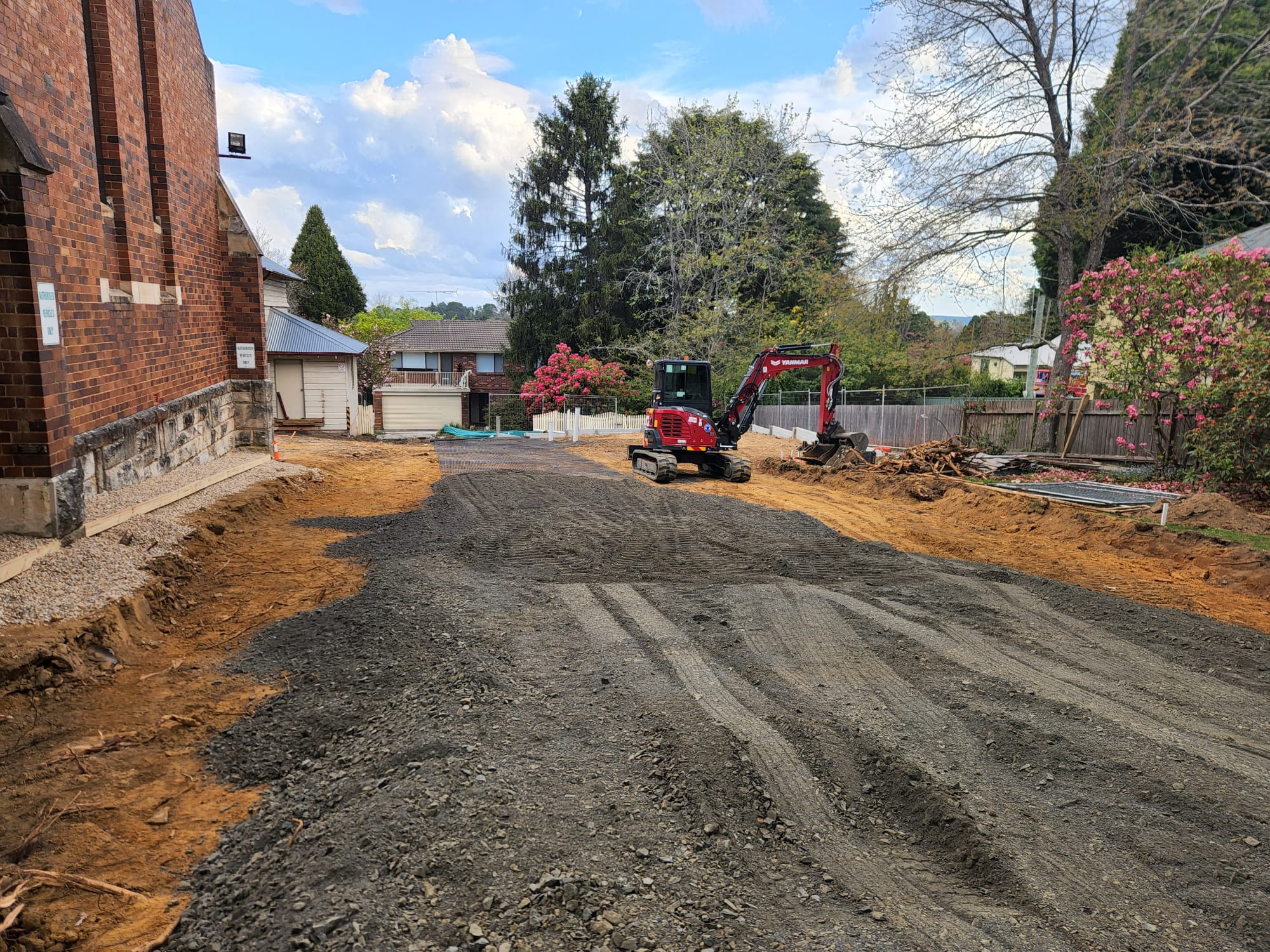 A construction site with a small red excavator on a gravel surface. The area is surrounded by trees and residential buildings, with clear blue skies above. There are piles of dirt and construction materials on the side.