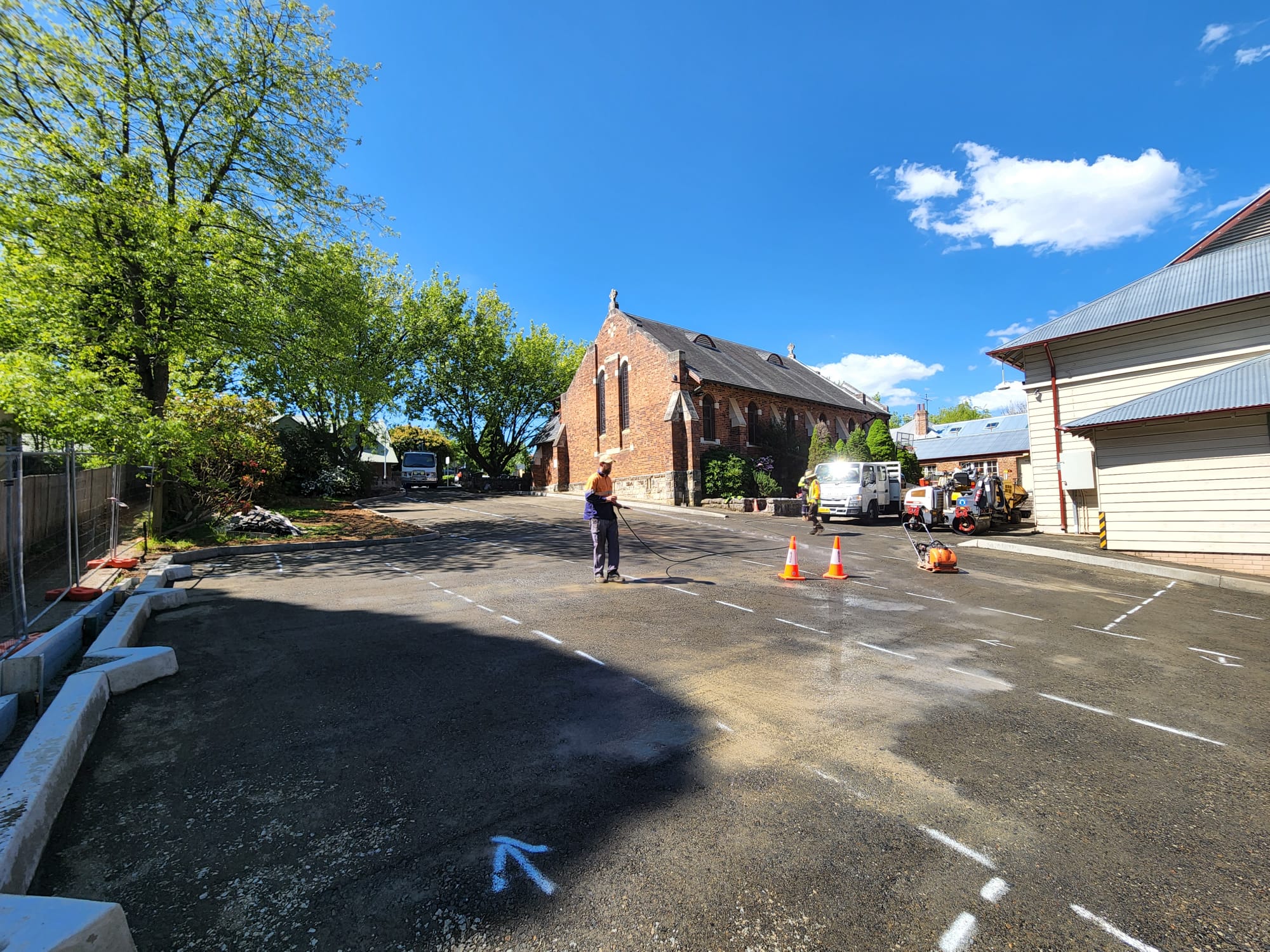 Person cleaning a paved driveway near a red brick building, with safety cones and vans in the background. A sunny day with blue sky and scattered clouds, surrounded by trees and buildings.