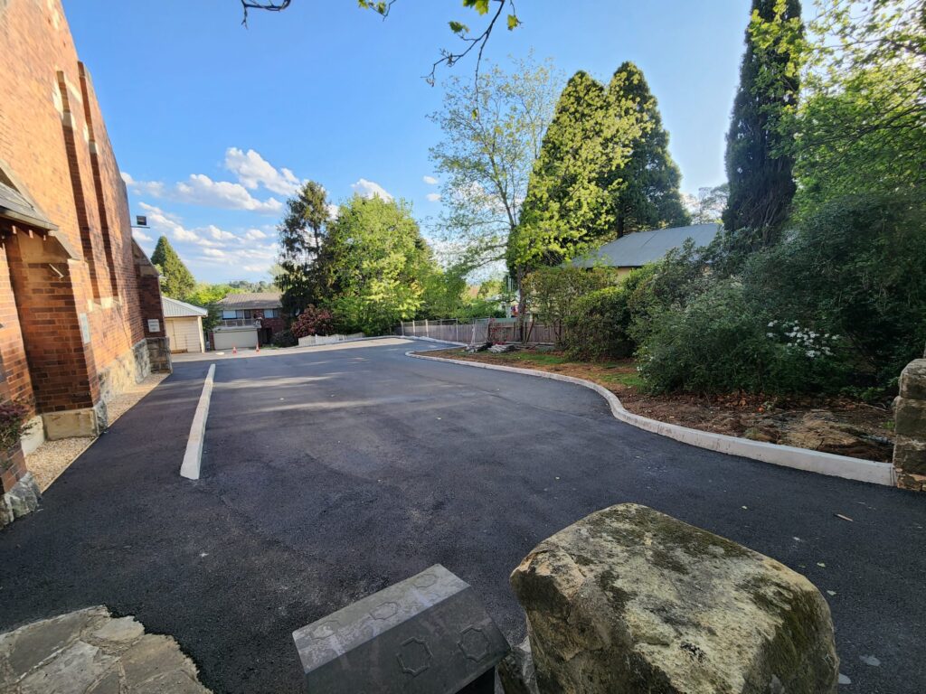 Paved driveway between a brick building on the left and a garden with trees on the right. The sky is clear and blue, with scattered clouds. A house is visible in the background. Stone curbs line the asphalt.